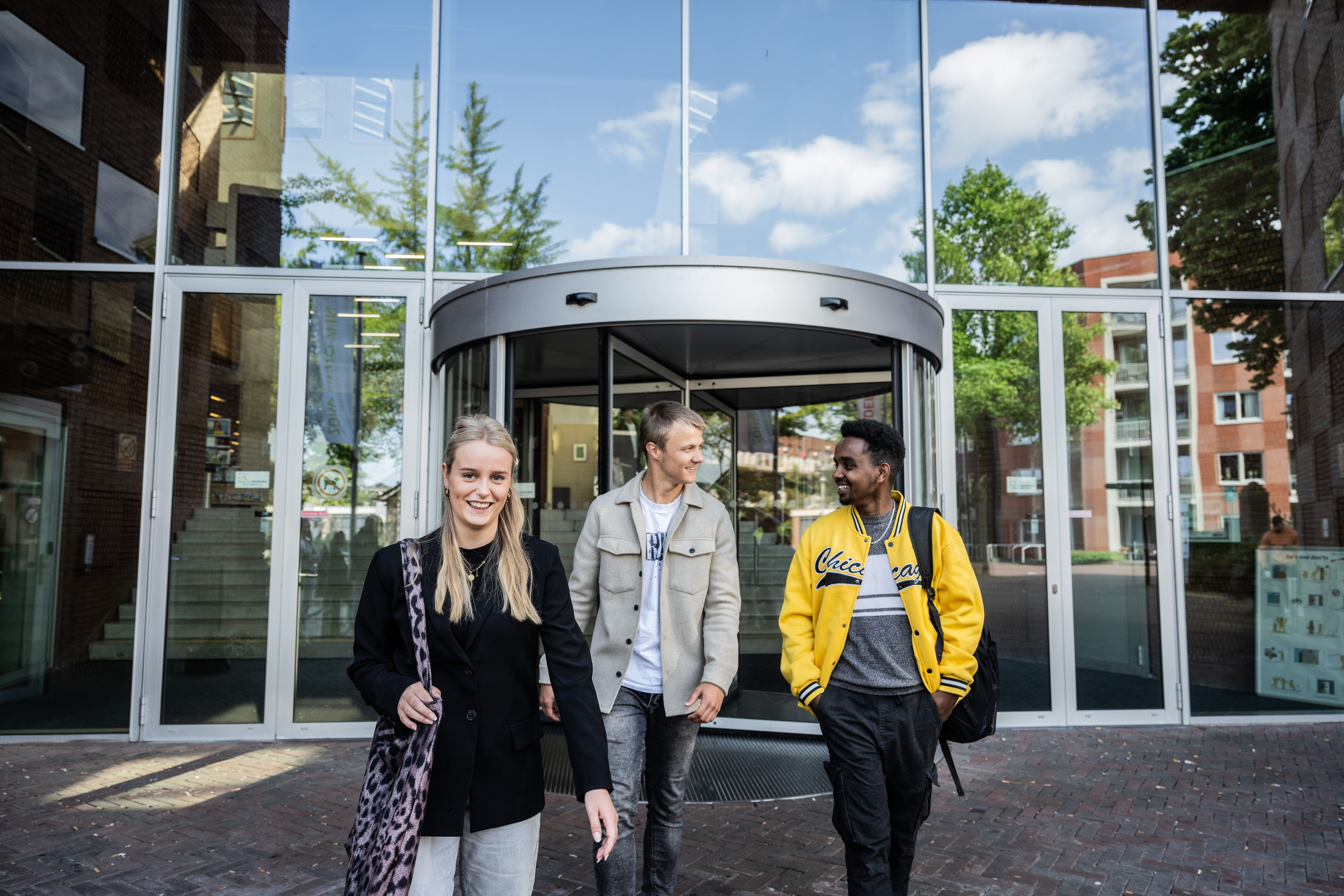Drie studenten verlaten een schoolgebouw met een glazen gevel.