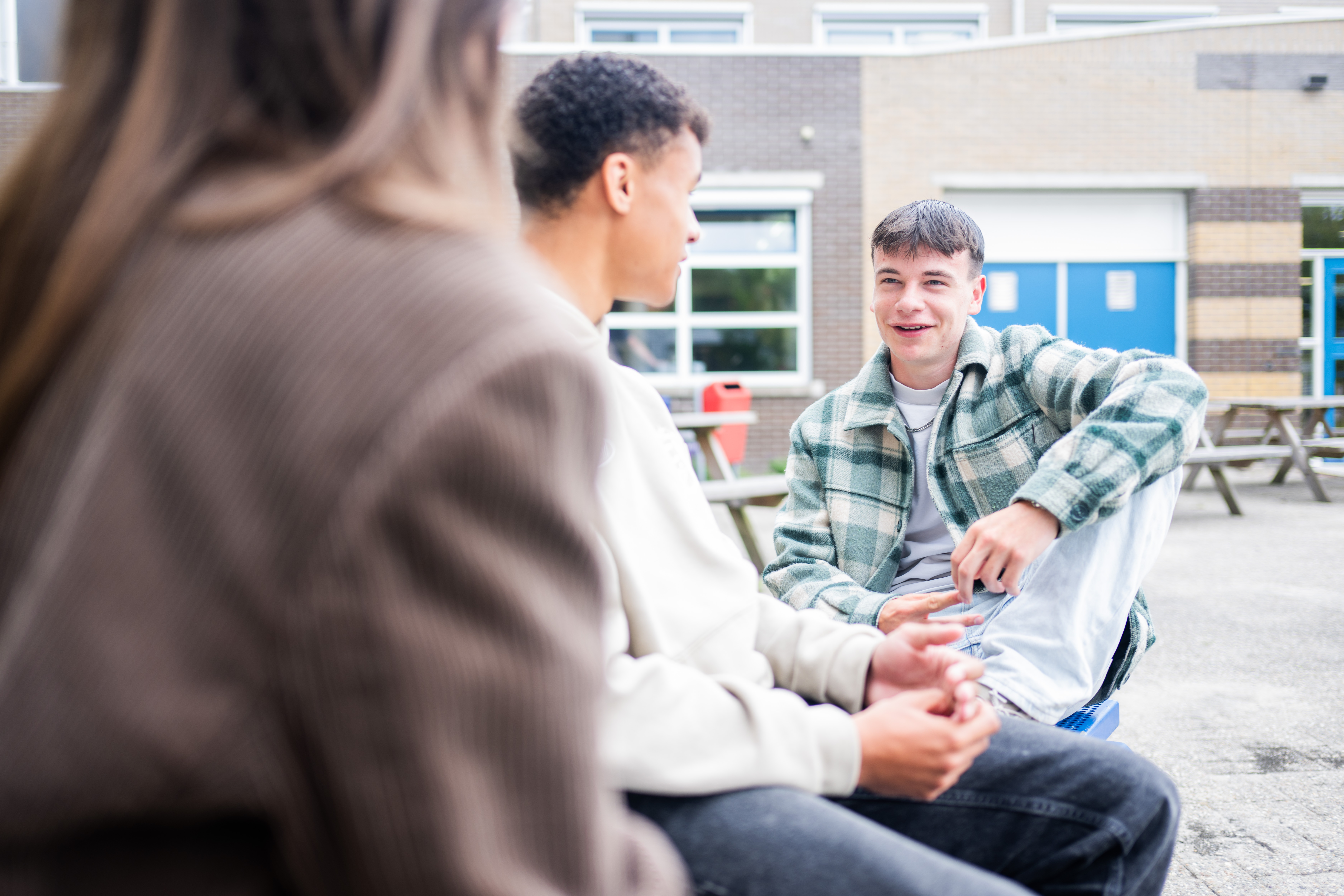 Drie studenten zitten buiten op het schoolterrein bij picknicktafels.