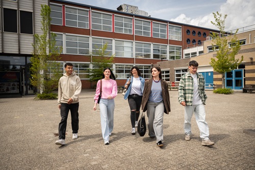 Vijf studenten lopen samen over het plein met het Alfa-college op de achtergrond.