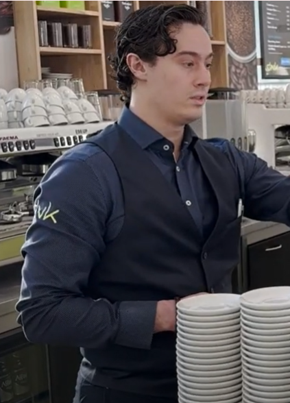 Student achter een balie in een café, met stapel borden en koffiemachines op de achtergrond.