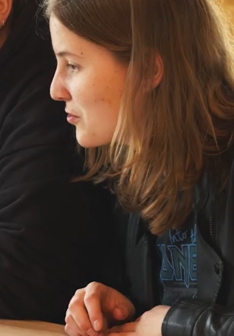 Student met lange haren en zwarte jas kijkt mee op laptop van medestudent.