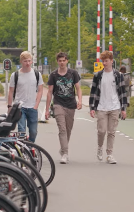 Drie studenten lopen op een pad langs geparkeerde fietsen en verkeersborden.