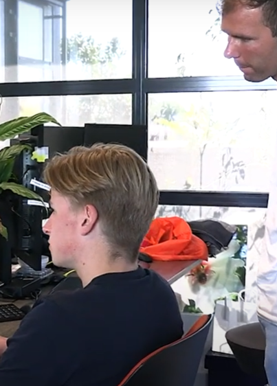 Twee personen in een kantoorruimte met bureau, computerschermen en uitzicht op bomen.