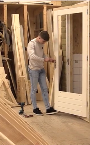 Student installeert een deur met glas in een werkplaats; gereedschap en houten planken zichtbaar.