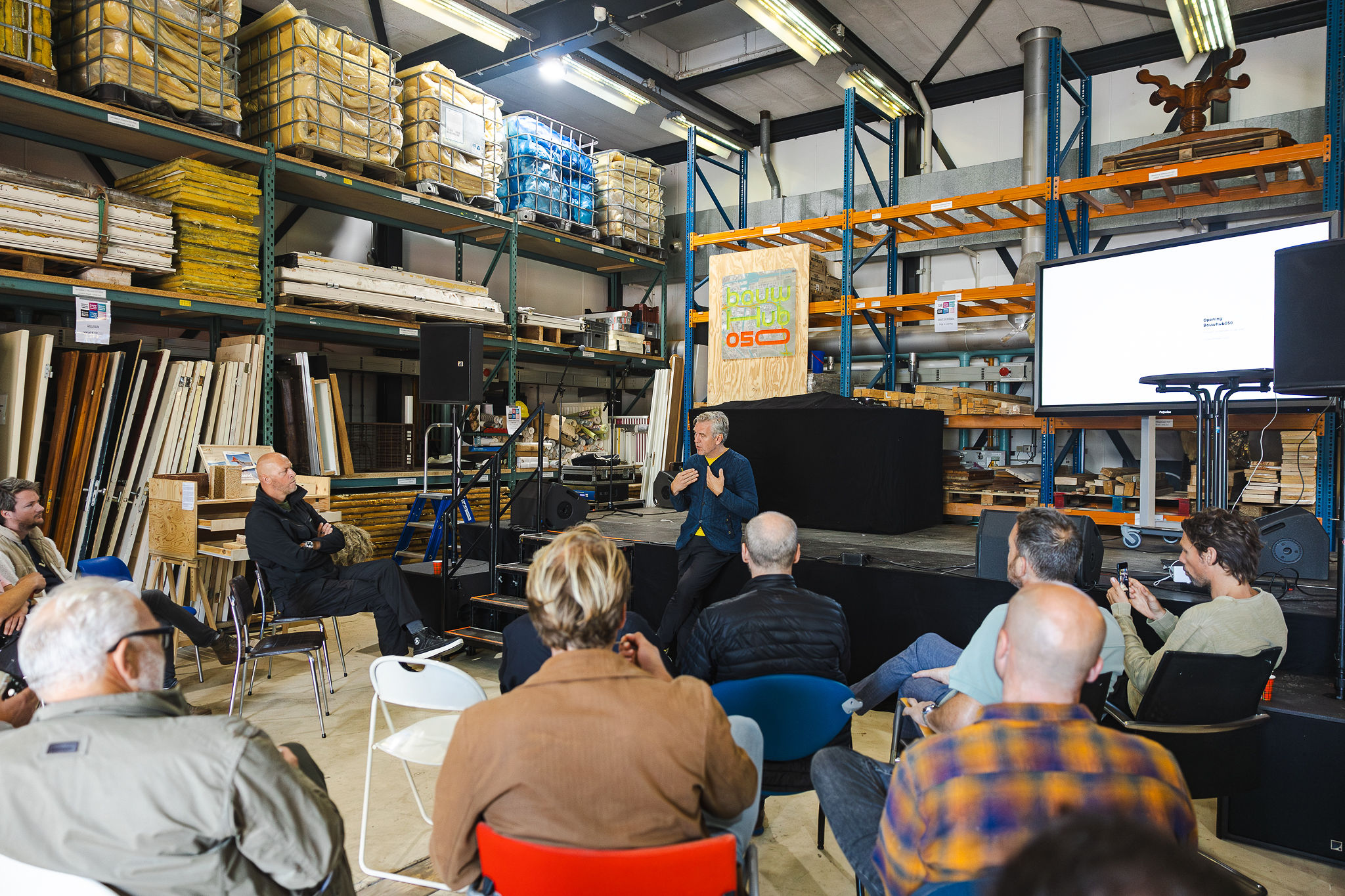 Mensen zitten op stoel te kijken naar een presentatie van een man in het gebouw van BouwHub050